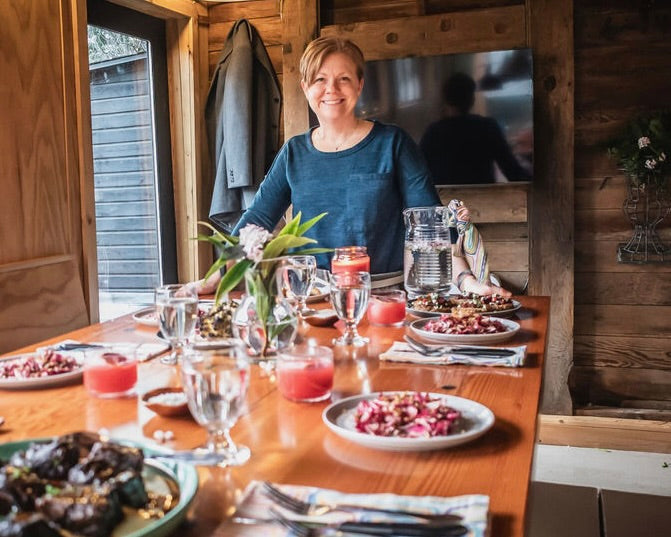 Woman sitting at a wooden table set for a meal with plates, glasses, and a pitcher.